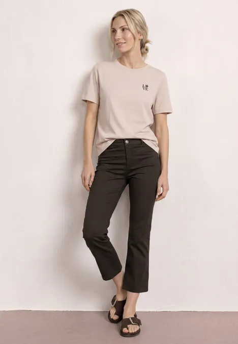 Frau in beigem T-Shirt, schwarzer 7/8-Hose und Sandalen vor neutralem Hintergrund.
