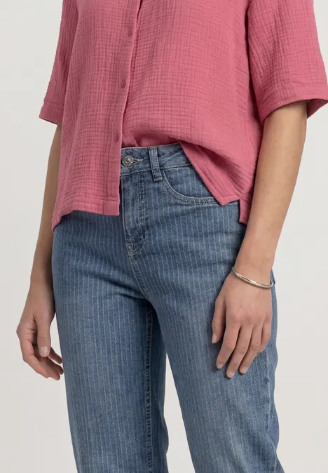 Een persoon draagt een roze blouse met korte mouwen, een blauwe jeans en een armband, alleen bovenlichaam en handen zichtbaar.