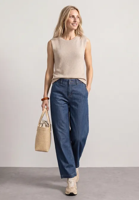 Een vrouw draagt een beige top, wijde blauwe jeans en sneakers, met een strooien tas voor een lichte achtergrond.