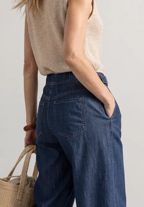 Vrouw van achteren in wijde jeans en beige top, ze draagt een strooien tas.