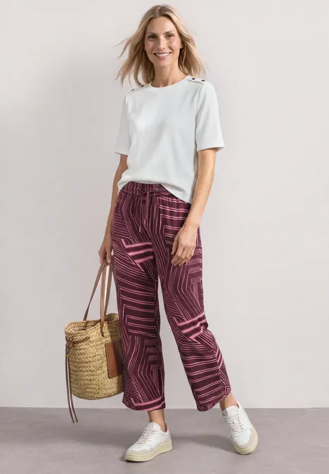 Femme en t-shirt blanc, pantalon violet à motifs, baskets blanches et sac en osier devant fond neutre.