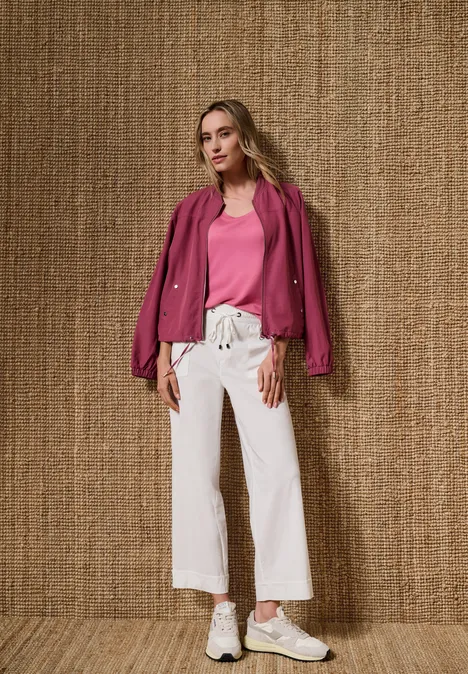 Vrouw in roze top en jasje, witte wijde broek en sneakers voor een textuurmuur.