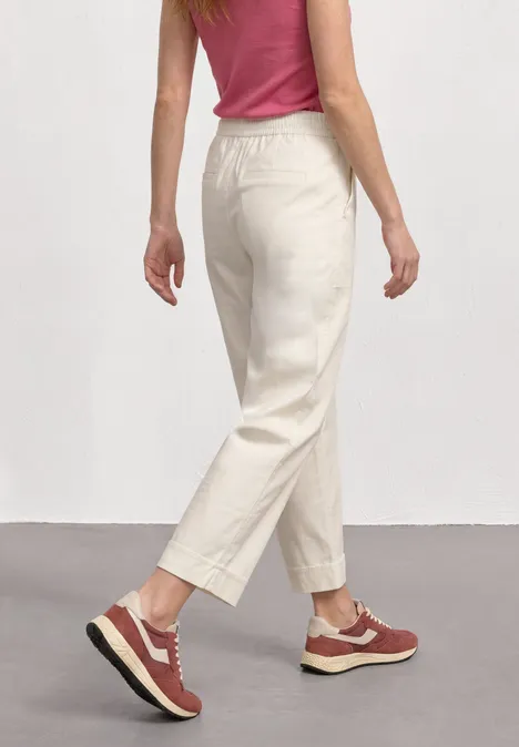 Vrouw van achteren in beige broek, roze top en rode sneakers tegen een lichtgrijze achtergrond.