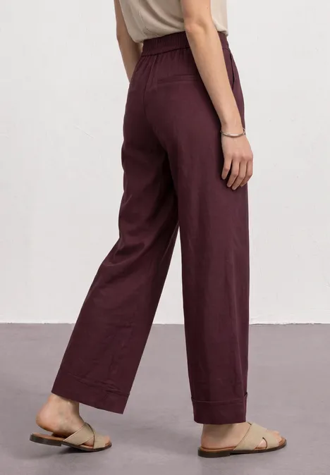Broek met Straight Leg pijpen in casual pasvorm en omgeslagen boorden rustic rouge Wijde, hoog getailleerde bordeaux linnen broek met platte sandalen voor een witte muur.