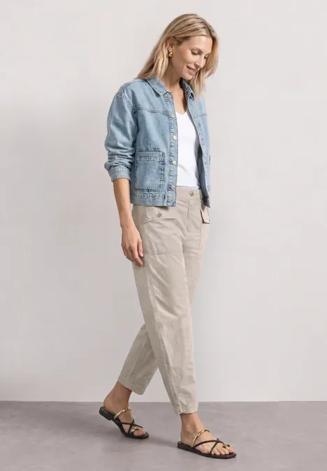 High Waist Barrel Leg Hose im Casual Fit clay sand Frau in hellblauer Jeansjacke, weißem Top und beigen Stoffhosen mit schwarzen Riemchensandalen vor hellem Hintergrund.