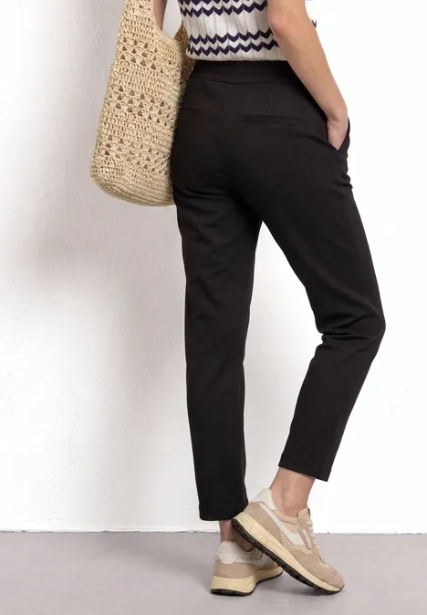 Vrouw van achteren in zwarte chino, gestreepte top, rieten tas en beige sneakers.
