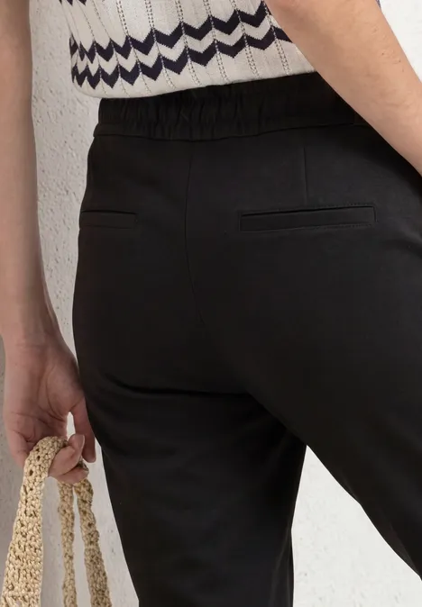 Detail van iemand in zwarte broek met elastische taille en achterzak, die een gehaakte tas vasthoudt.