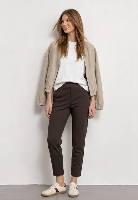 Een jonge vrouw in wit shirt, beige blazer, donkere broek en sneakers, poseert voor een lichte achtergrond.