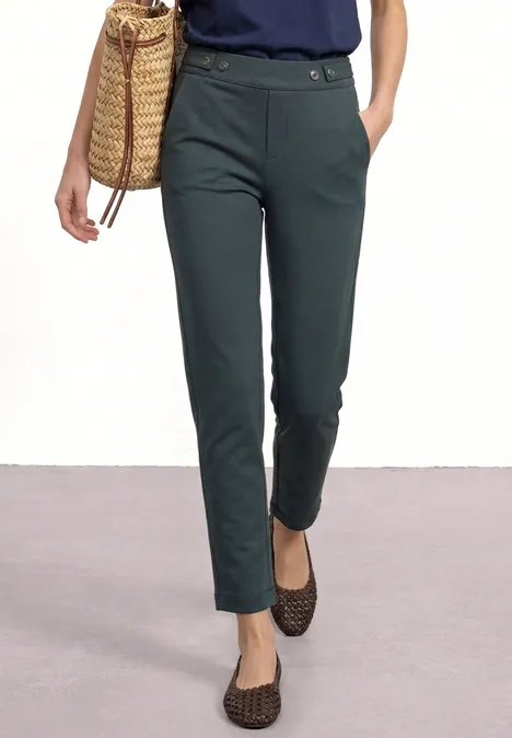 Vrouw draagt een olijfgroen, smal pantalon met sierceintuur, gecombineerd met gevlekte ballerina’s en een rieten tas.