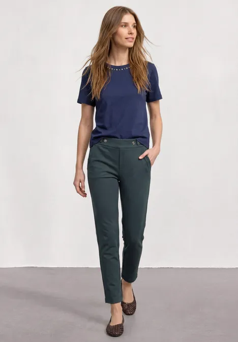 Vrouw in smalle donkergroene broek, marineblauw T-shirt en bruine schoenen op lichte achtergrond.