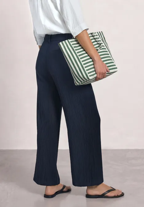 Frau mit weiten dunkelblauen Hosen und schwarzen Flip-Flops, hält eine grün-beige gestreifte Tasche.
