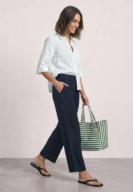 Eine Frau in weißem Hemd, blauer Culotte, Flipflops und gestreifter Tasche, im Profil gehend.