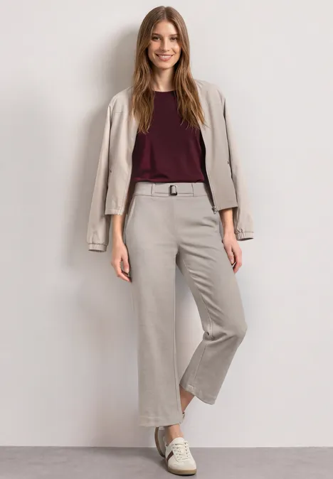 Vrouw in beige cropped broek met bijpassend jasje over de schouders, bordeaux top en witte sneakers.