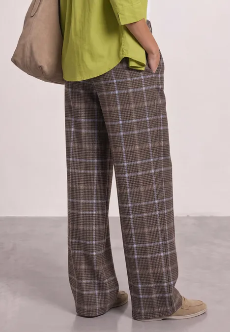 Hose mit braunem Karomuster, weites Bein, kombiniert mit grünem Overshirt, Beuteltasche und beigen Schuhen.