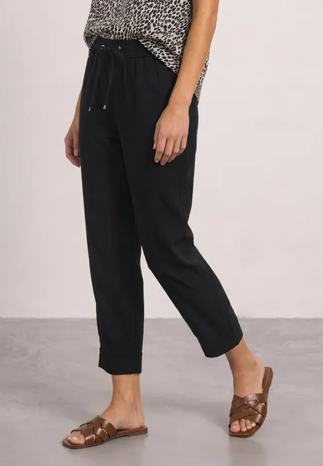 Vrouw draagt zwarte, enkellange broek met losse pasvorm en bruine, platte sandalen.