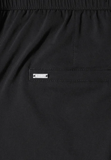 Close-up van een zwarte broek met elastische tailleband en een klein metalen plaatje bij de zak.