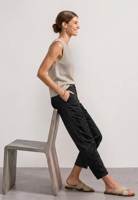 Femme de profil, tenue décontractée: top sans manches, pantalon noir, sandales, appuyée sur un tabouret en bois.