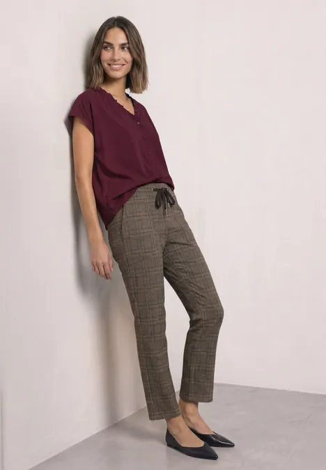 Mid Waist Slim Leg Hose im Loose Fit saddle brown Eine Frau lehnt an einer Wand, trägt bordeauxrotes Oberteil, karierte Hose und schwarze Ballerinas.