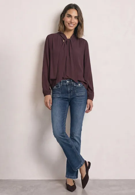 Frau in bordeauxrotem Blusenshirt und blauen Bootcut-Jeans steht lächelnd vor hellem Hintergrund.