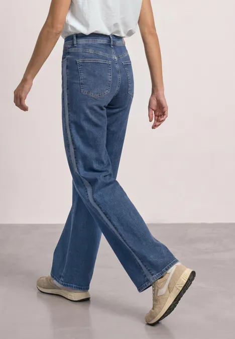 Rückansicht einer Person in weitem blauen Jeans und hellen Schuhen vor neutralem Hintergrund.