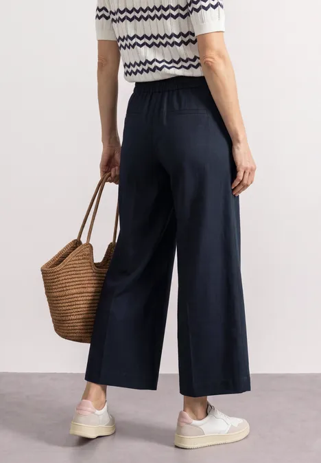 Persoon draagt marineblauwe culotte, gestreept topje, witte sneakers en een rieten tas.