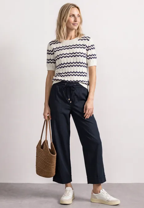 Een vrouw draagt een gestreept shirt, wijdvallende broek, witte sneakers en houdt een gevlochten tas vast.