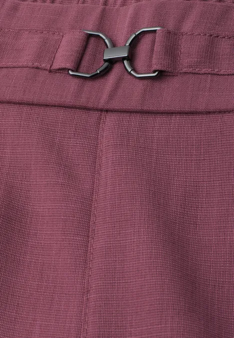 Gros plan d’un pantalon couleur prunelle à la texture délicate, agrémenté d’une boucle métallique argentée.