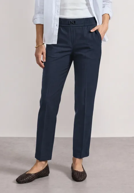Slim Leg Hose mit Schnallendetail darkest blue Dame in dunkelblauen Chino-Hosen mit Bügelfalte trägt weiße Bluse und geflochtene schwarze Loafers.