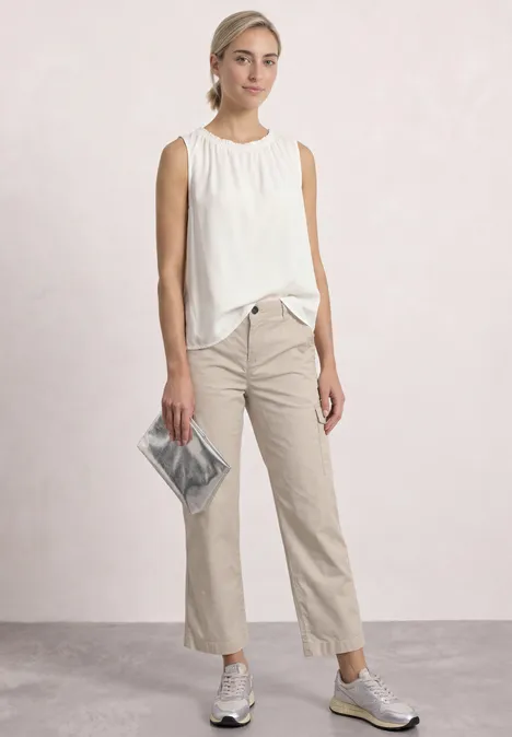 Femme en haut blanc sans manches, pantalon beige, baskets blanches et pochette argentée sur fond clair.