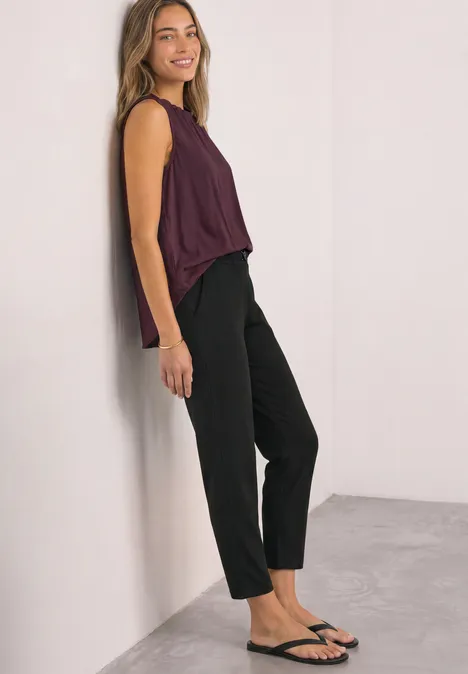 Slim Leg Hose mit Schnallendetail Black Frau lehnt an Wand, trägt bordeauxfarbenes Top, schwarze Hose und Sandalen, lächelt leicht.