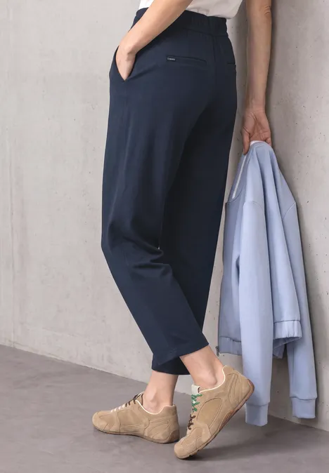 Pantalon Slim Leg à détails boucles darkest blue Damenbein in dunkelblauer Hose, hält hellblaue Jacke, trägt beigen Sneaker.