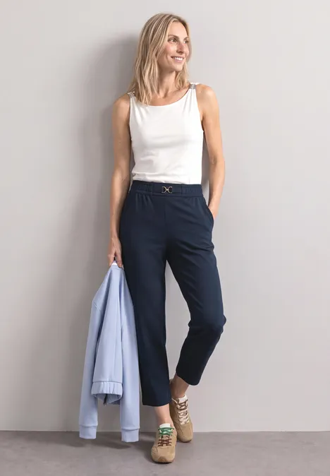 Pantalon Slim Leg à détails boucles darkest blue Eine blonde Frau lehnt an der Wand, trägt weißen Top, dunkelblaue Hose und Espadrilles, hält hellblauen Blazer.