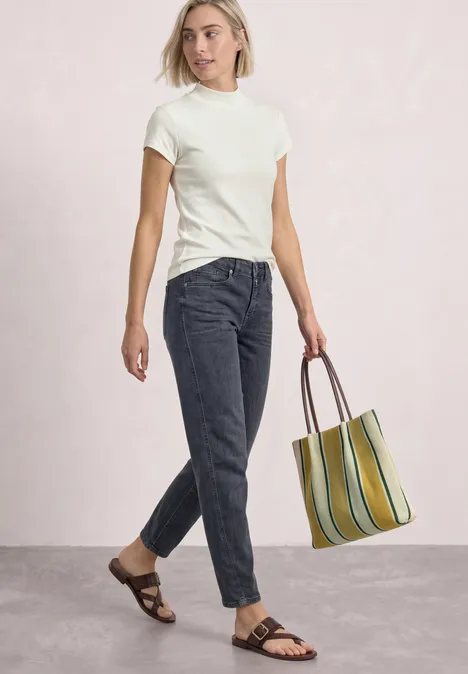 High Waist Barrel Leg Jeans im Loose Fit Steel Grey Washed Frau in weißem T-Shirt, dunkler Jeans, Sandalen und gestreifter Tragetasche vor hellem Hintergrund.