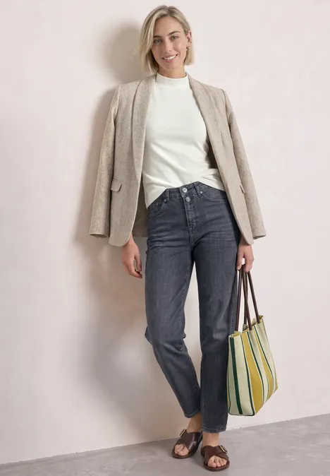 High Waist Barrel Leg Jeans im Loose Fit Steel Grey Washed Frau in weißem Pullover, beigem Blazer, dunklen Jeans, Sandalen und gestreifter Tasche vor neutralem Hintergrund.