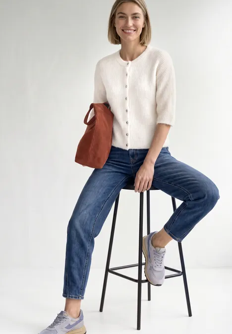 High Waist Barrel Leg Jeans im Loose Fit Mid Blue Random Washed Frau sitzt auf Hocker, trägt cremefarbenen Cardigan, blaue Jeans, weiße Sneaker und hält eine braune Tasche.