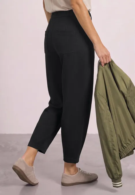 Frau trägt schwarze, hoch geschnittene, schmal zulaufende Hose, beige Sneaker und olivgrüne Jacke.