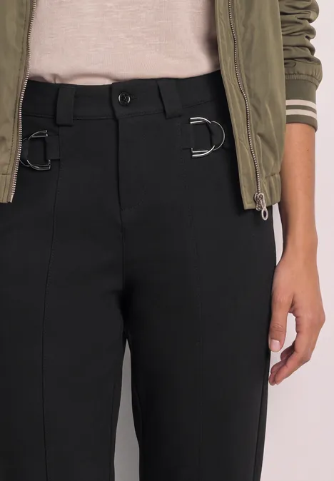 Schwarze Hose mit D-Ringen an der Taille, kombiniert mit beigem Shirt und grüner Jacke.