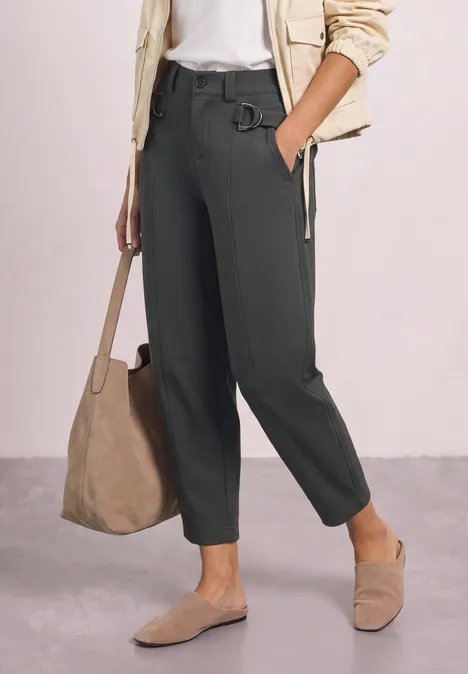 Frau trägt hoch taillierte dunkelgrüne Hose mit Gürtel, weißes Top, beige Jacke, Slipper und Wildledertasche.