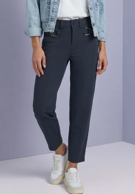 Schwarze 7/8-Hose und weiße Sneaker, kombiniert mit einer Jeansjacke vor fliederfarbenem Hintergrund.
