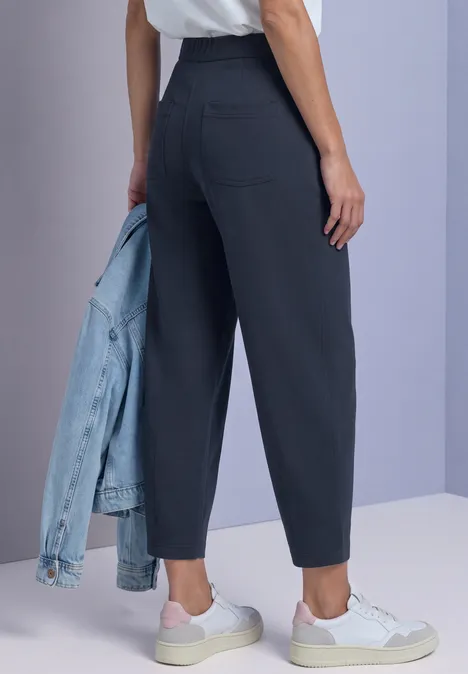 Frau in dunkelgrauen High-Waist-Hosen, weißen Sneakern und hält eine Jeansjacke vor hellem Hintergrund.