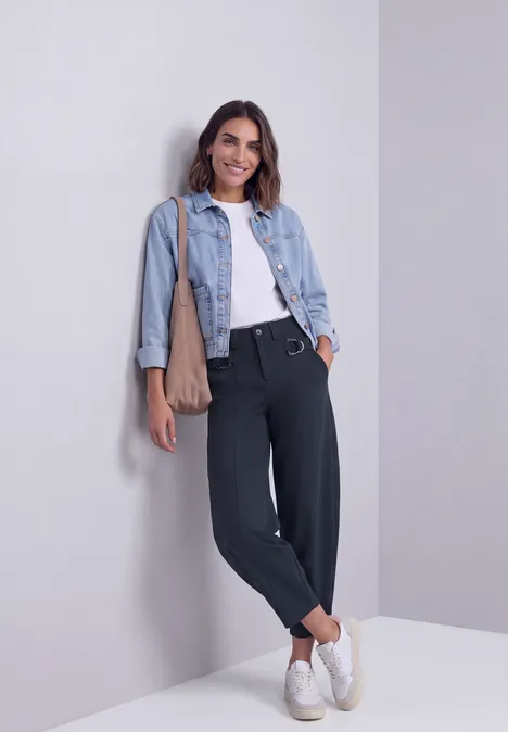 Junge Frau lehnt lässig an einer Wand, trägt Jeansjacke, weißes Top, schwarze Hose und weiße Sneaker.