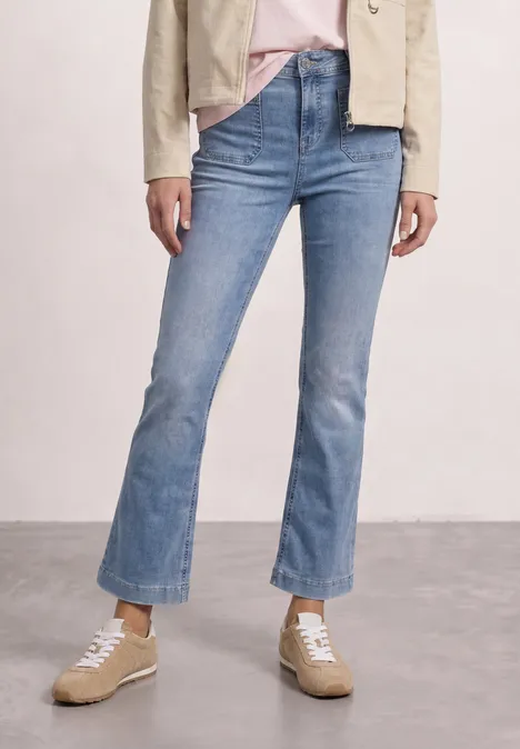 Vrouw draagt lichtblauwe uitlopende jeans met ruwe zoom, beige sneakers, roze shirt en beige jasje.