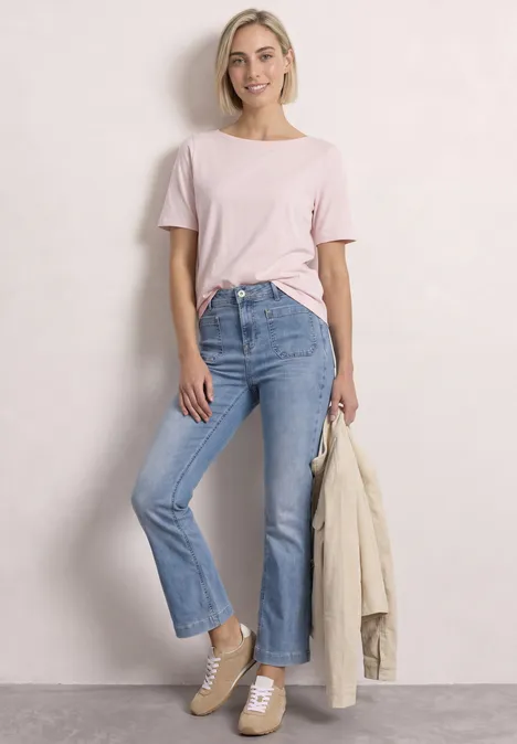 Jong blond vrouwtje in roze T-shirt, lichtblauwe jeans en beige sneakers, met een beige jas in de hand.