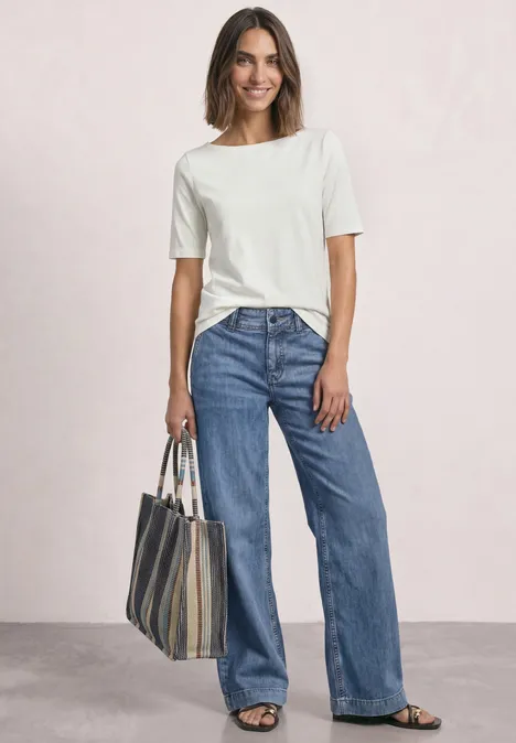 Vrouw in wit T-shirt en losse jeans, met gestreepte tas, glimlachend voor een lichte achtergrond.