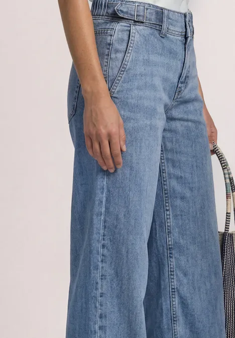 Vrouw draagt een wijde blauwe spijkerbroek en houdt een gevlochten tas vast.
