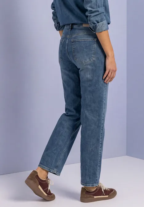 Rückansicht einer Person in blauen Jeans und braunen Schuhen vor einfarbigem Hintergrund.
