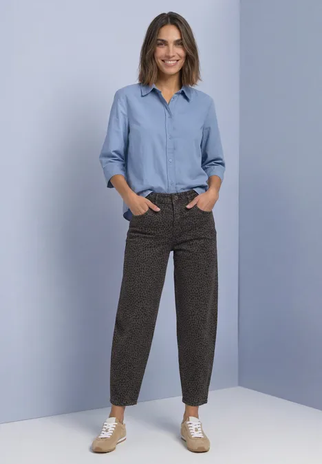 High Waist Barrel Leg Jeans im Loose Fit brown leo washed Eine lächelnde Frau in hellblauer Bluse und braunen, gemusterten Hosen mit Sneakers vor hellblauem Hintergrund.