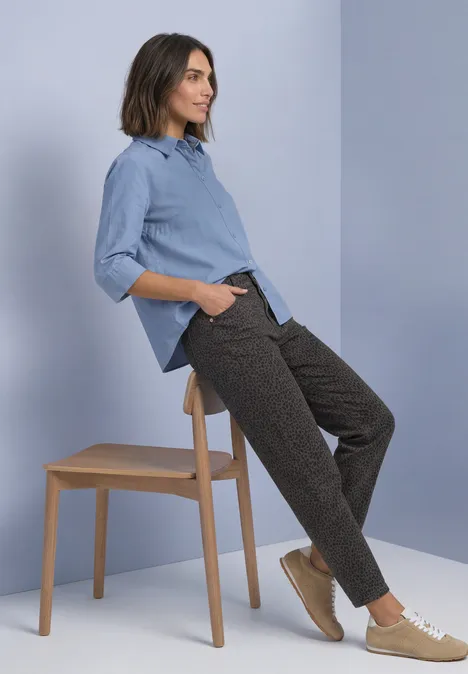 High Waist Barrel Leg Jeans im Loose Fit brown leo washed Frau stützt sich lässig an Hocker ab, trägt hellblaues Hemd, dunkle Hose und beige Sneaker.