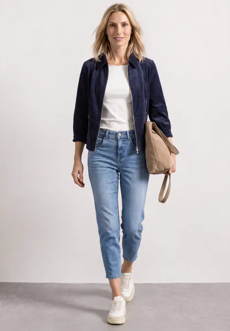 Een vrouw in wit shirt, donkerblauw jasje en jeans draagt een tas en loopt glimlachend.