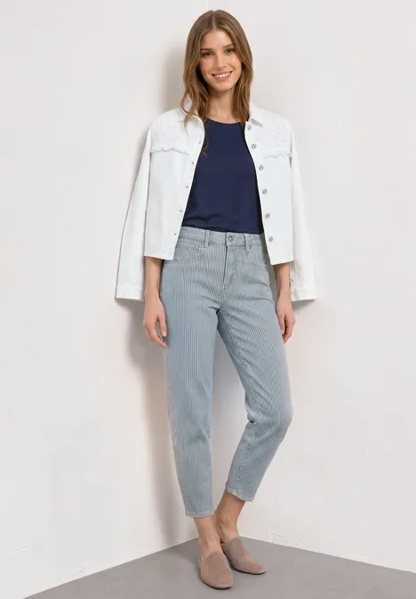 Femme en veste blanche sur t-shirt bleu marine et pantalon gris clair, debout devant un mur clair.